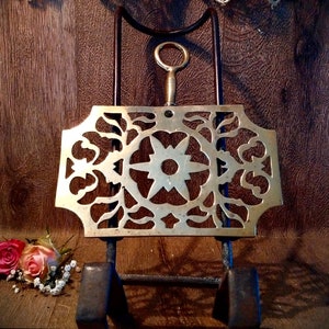 Może przedstawiać: Metalowy stojak na tosty z mosiądzu z ozdobnym wzorem kwiatowym. Stojak jest zamontowany na czarnym metalowym stojaku.