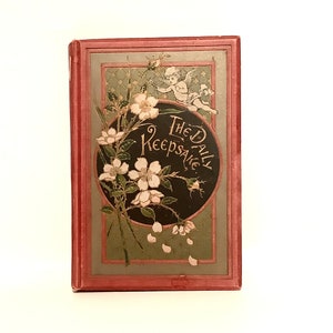 Puede incluir: La portada de un libro antiguo titulado "The Daily Keepsake" con un diseño floral con flores blancas y un querubín. La portada es roja con un círculo negro en el centro.