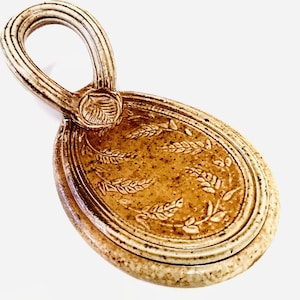 May include: A handmade ceramic soap dish with a handle. The oval dish is light brown with a textured surface and a raised leaf design. The handle is a loop shape with a ribbed texture.