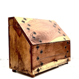 Op de afbeelding: Een houten doos met een scharnierend deksel en een kleine lade. De doos is versierd met metalen accenten en heeft een natuurlijke houten afwerking.