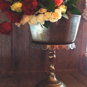 May include: A silver pedestal bowl with a scalloped edge sits on a gold stand. The bowl is filled with a bouquet of red, white, and yellow roses.