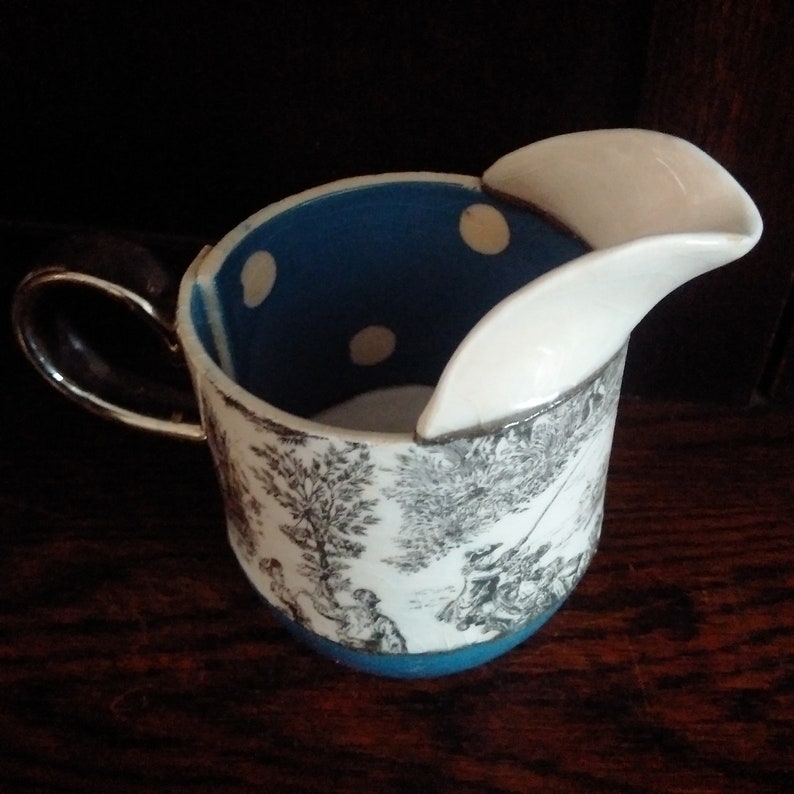 May include: A white ceramic creamer with a blue and white polka dot interior and a black handle. The creamer has a unique spout that is separate from the main body.