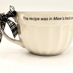 Pode incluir: Tigela de mistura de cerâmica branca com uma fita xadrez preta e branca amarrada à alça. A tigela tem uma borda ondulada e o texto "The recipe was in Maw's heid and the" está impresso no lado.