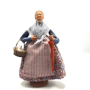 May include: A ceramic figurine of an older woman wearing a blue and white patterned dress with a pink and white striped apron. She is holding a basket with a white bird and an orange umbrella.