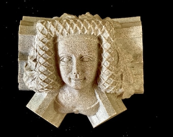 May include: A stone sculpture of a woman's head with a braided hairstyle. The sculpture is carved in a detailed style and has a weathered finish.