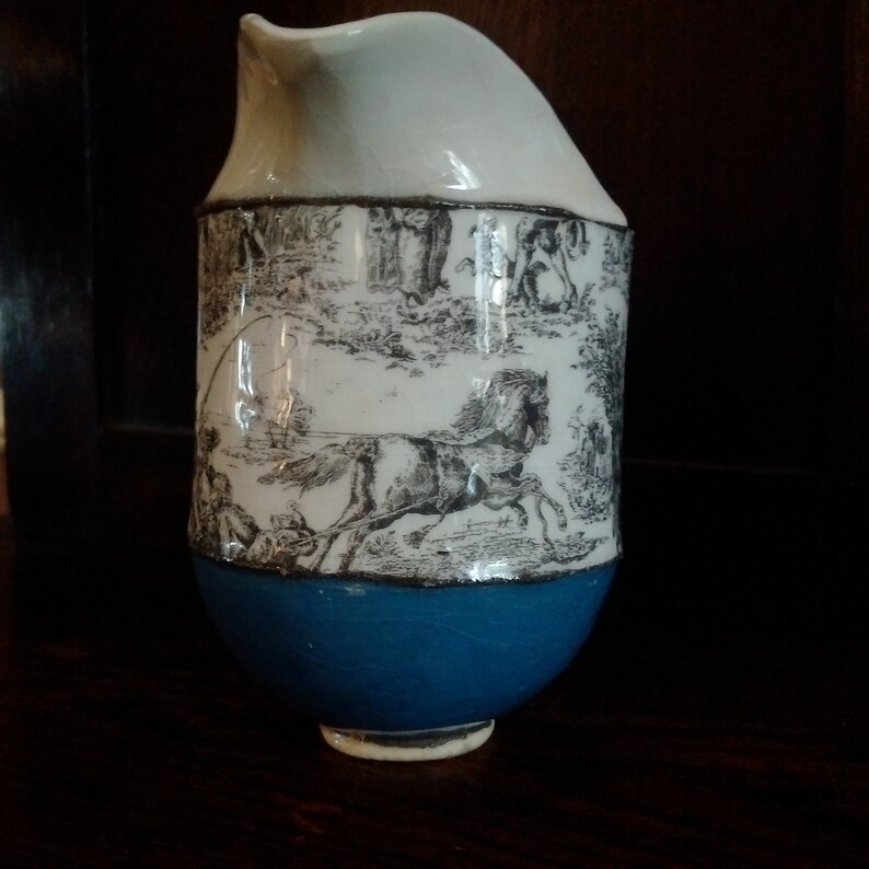 May include: A white ceramic pitcher with a blue base. The pitcher has a black and white design of a horse and rider.