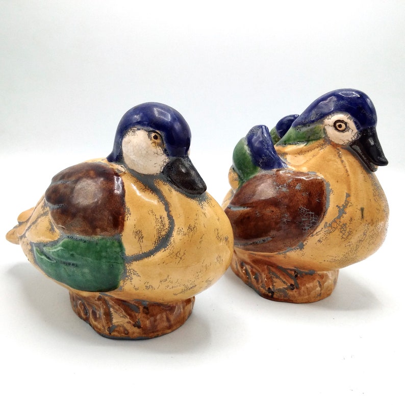 Pu&ograve; includere: Due statuine di anatra in ceramica dipinte in marrone, verde e blu. Le anatre sono rivolte in direzioni diverse e hanno un aspetto usurato.