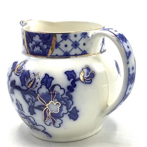 Puede incluir: Una jarra de crema de cerámica blanca con un diseño floral azul y dorado. La jarra tiene un cuerpo redondo y un asa.