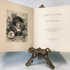 May include: An open book titled "Lady Clare" by Alfred Tennyson, with an illustration of a woman in a hat surrounded by roses. The book is held open by a decorative brass stand with ornate detailing.