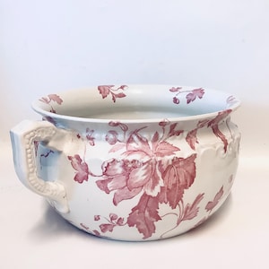 Può includere: Vaso da notte in ceramica bianca con un grande motivo floreale nei toni del rosa. Il vaso ha un manico su un lato e un bordo largo. Il motivo floreale comprende foglie e fiori.