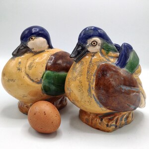 Pu&ograve; includere: Due statuine di anatra in ceramica con smalto blu, marrone e verde. Le anatre sono sedute su una superficie bianca con un uovo marrone davanti a loro.