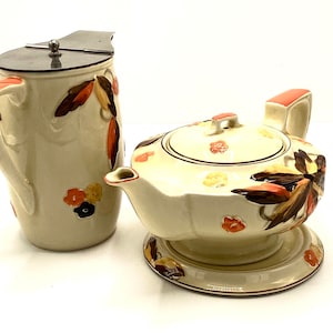 Puede incluir: Un juego vintage de tetera y jarra de cerámica de color crema con un patrón floral marrón, naranja y amarillo. La tetera tiene una tapa y una base a juego. La jarra tiene una tapa de plata.