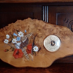 May include: A wooden wall decor featuring a painted scene of a bird perched on a branch with wildflowers, including poppies, daisies, and bluebells. A round, silver-toned barometer with a black needle is also painted on the wood.