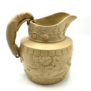 May include: A beige ceramic jug with a sculpted dog handle. The jug features a relief design of a hunting scene with trees and hounds.