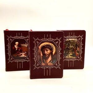 Op de afbeelding: Drie kleine bordeauxrode lederen boeken met gouden accenten en religieuze afbeeldingen op de omslagen. De omslagen tonen afbeeldingen van Jezus, een vrouw met een baby en een scène met mensen die lopen.