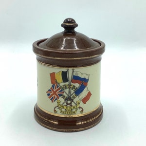 May include: A brown ceramic jar with a lid, featuring a design of four flags crossed together. The flags are the British Union Jack, the Belgian flag, the Russian flag, and the French flag. The jar has a gold trim and the text "FOR RIGHT AND FREEDOM" around the bottom.