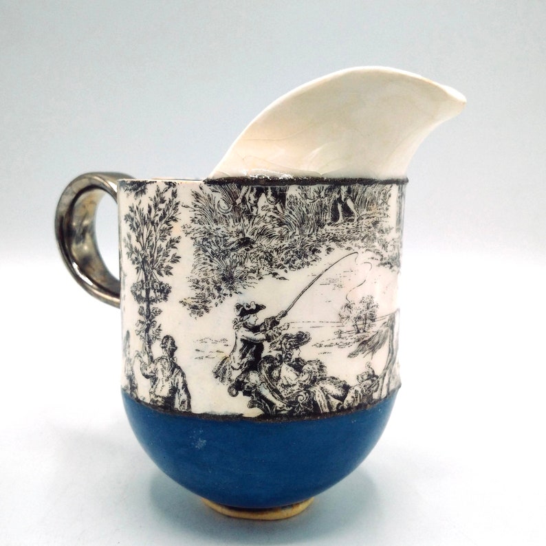 May include: A white ceramic creamer with a blue base and a black and white print of a fishing scene. The creamer has a unique spout design.