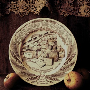 May include: A vintage ceramic plate with a brown and white design featuring a variety of breads and cheeses. The plate has the text "British Bread and Cheese Plate" printed on it. The text "Caerphilly Cheddar Cheshire Gloucester Stilton Wensleydale" is printed around the bottom of the plate. The text "Oats Wheat Rye-Barley" is printed in the center of the plate.