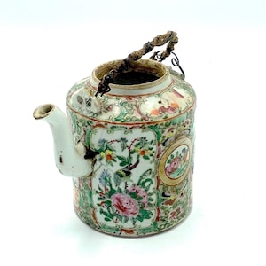 May include: A small, white porcelain teapot with a floral design and a brown woven handle. The teapot has a spout and a lid.