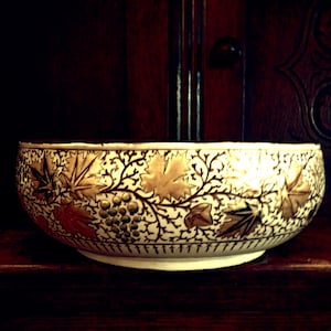 Pode incluir: Uma tigela de cerâmica branca com um design de videira dourado e marrom. A tigela é redonda e tem uma borda larga.