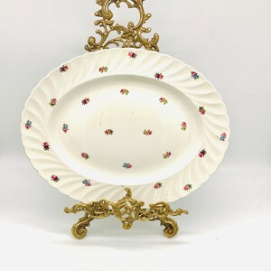 May include: A white oval porcelain plate with a floral pattern, displayed on a gold metal stand. The plate has a raised edge and a scalloped design.
