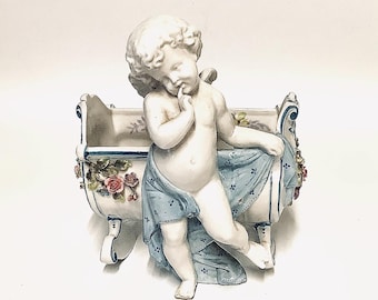 Puede incluir: Una figurita de porcelana de un querubín de pie en una maceta blanca y azul con detalles florales. El querubín sostiene un paño azul y tiene una expresión pensativa.