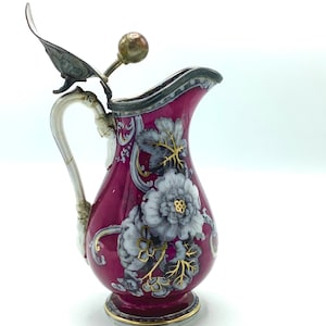 May include: A vintage porcelain pitcher with a floral design in shades of pink, white, and gold. The pitcher has a silver lid with a decorative knob.