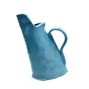 Puede incluir: Una jarra de cerámica azul claro con una forma única y asimétrica. La jarra presenta un diseño decorativo de flores en relieve y patrones geométricos.