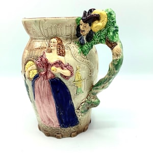 May include: A ceramic pitcher with a whimsical design featuring a woman in a pink and blue dress, a man with a green hat, and a leafy handle. The pitcher has a white background with brown, green, and yellow accents.