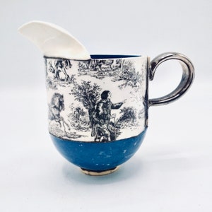 May include: A white ceramic creamer with a black and white toile pattern featuring a man with a walking stick and a horse. The creamer has a blue base and a silver handle.
