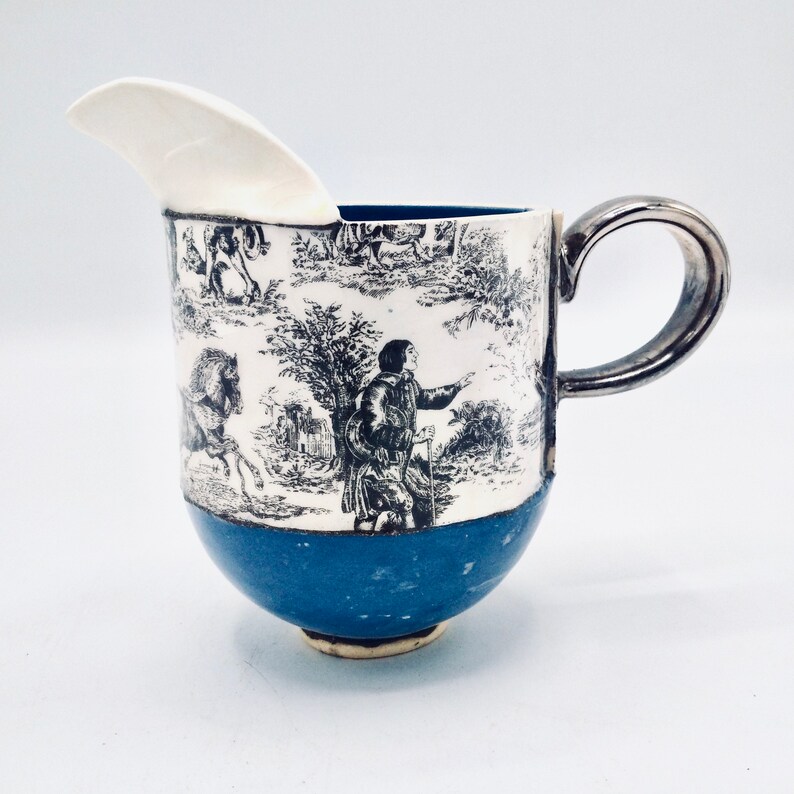 May include: A white ceramic creamer with a black and white toile pattern featuring a man with a walking stick and a horse. The creamer has a blue base and a silver handle.