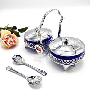 Könnte beinhalten: Ein silbernes Zucker- und Sahne-Set mit blauen Glasschalen und einem dekorativen Gitterdesign. Das Set enthält zwei Löffel mit einem muschelförmigen Design.