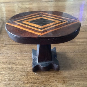 Può includere: Un piccolo tavolo in legno con un intarsio a forma di diamante di legni di diversi colori. Il tavolo ha un piano arrotondato e una base a piedistallo.
