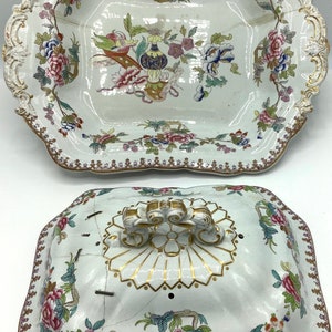 May include: A white porcelain serving dish with a lid. The dish and lid are decorated with a floral pattern in shades of pink, blue, and green. The lid has a gold handle and a gold design in the centre.
