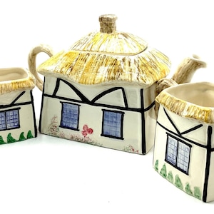 May include: A white ceramic teapot and two cups shaped like cottages with thatched roofs. The teapot has a brown thatched roof, white walls, and blue windows. The cups have brown thatched roofs, white walls, and blue windows. The cottages have green grass and flowers painted on the sides.