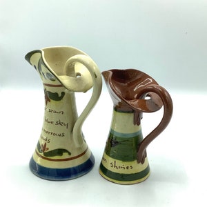 Puede incluir: Dos jarras de cerámica con un diseño único y caprichoso. La jarra de la izquierda es blanca con detalles azules y verdes y el texto "the sun blue sky tomorrows clouds". La jarra de la derecha es marrón con detalles verdes y amarillos y el texto "sun shines".