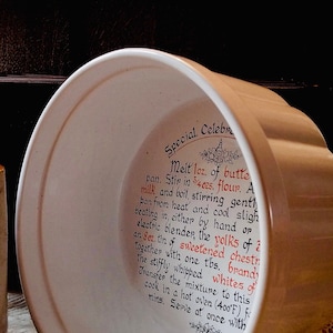 May include: White ceramic baking dish with a scalloped rim. The inside of the dish has handwritten instructions for a special celebration cake recipe, including ingredients like butter, flour, yolks, and brandy.