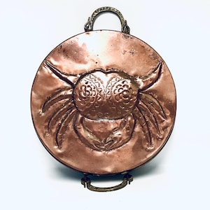 May include: A round copper wall hanging featuring an embossed design of a crab. The hanging has a brass handle and a brass stand.