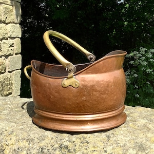 Può includere: Un secchio per carbone in rame vintage con manico in ottone e dettagli decorativi. Il secchio è appoggiato su una superficie di pietra.
