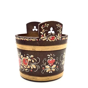 May include: A wooden painted container with a brown base and floral designs in red, yellow, and white. The container has two handles on the top and a rounded shape.