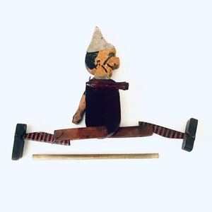 May include: A wooden toy figure of a person with a white hat, painted brown, black, and white, with a stick attached to the figure's legs. The figure is in a split position with the legs extended out to the sides. The figure is on a white background.