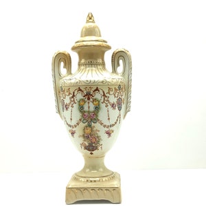 May include: A white ceramic vase with gold accents and floral designs. The vase has two handles and a lid. It sits on a square base.