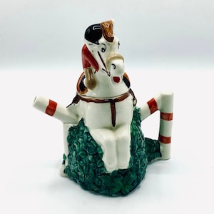 Könnte beinhalten: Eine weiße Keramik-Teekanne in Form eines Pferdes mit einem Reiter. Das Pferd sitzt auf einem grünen Sockel mit Blättern. Die Teekanne hat einen rot-weiß gestreiften Griff.