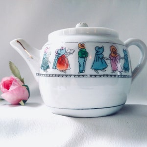 May include: A white porcelain teapot with a gold handle and a whimsical design of children in colorful clothing. The teapot has a lid with a knob on top.
