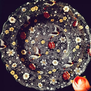 Puede incluir: Un plato decorativo con un fondo oscuro y un detallado patrón floral y de pájaros. El plato presenta flores blancas, amarillas y rojas, junto con pájaros en vuelo. El plato tiene un borde festoneado.