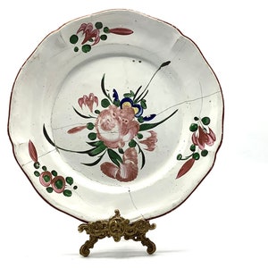 May include: An antique white plate featuring a floral design in pink, green, and blue. The plate has a scalloped edge with a red border and visible cracks. It is displayed on a decorative brass stand.