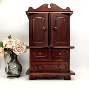 Può includere: Un mobiletto in legno scuro con due ante e tre cassetti. Il mobiletto ha una parte superiore decorativa e poggia su quattro gambe.