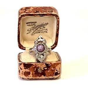 May include: A silver ring with a purple stone set in a decorative design. The ring is in a small brown box with the text "Watchmaker Jeweller J. Carruthers 59 High St. Skipton Telephone 222" on the inside.