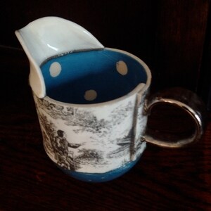 May include: A white ceramic pitcher with a blue and white polka dot interior and a black and white landscape design. The pitcher has a unique spout and a silver handle.