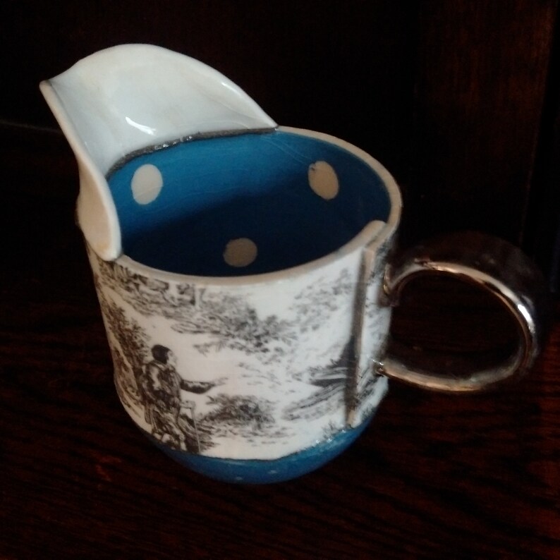 May include: A white ceramic pitcher with a blue and white polka dot interior and a black and white landscape design. The pitcher has a unique spout and a silver handle.